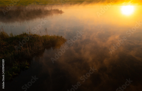 Spring landscape with the morning fog rising above a beautiful lake next to a rapeseed field. Agriculture in amazing parts of the world. Foggy aerial view during sunrise.