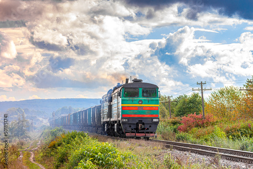 A powerful green diesel locomotive pulls a long train loaded with cement and building materials. Freight railway transportation. Sunny weather.