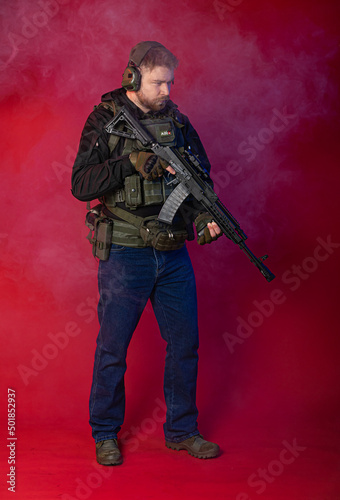 airsoft player in full gear with guns GG RK74. a man in headphones, body armor, with a backpack and a belt. isolated, red background. in blue smoke © Ольга Новицкая