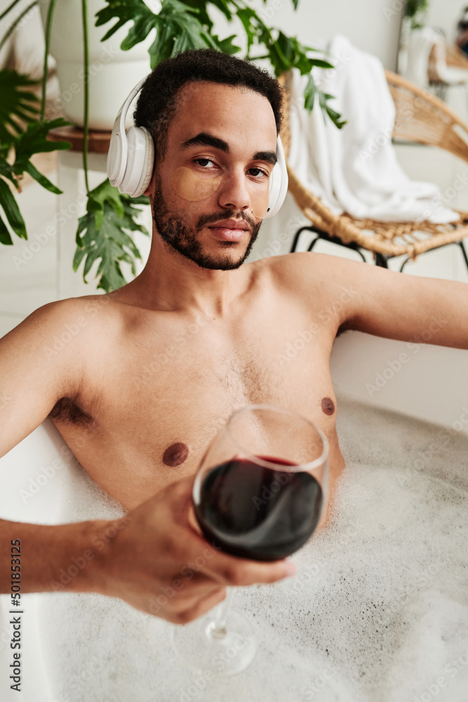 Portrait of young man in wireless headphones looking at camera while drinking wine and taking a bath Portrait of young man in wireless headphones looking at camera while drinking wine and taking a bath