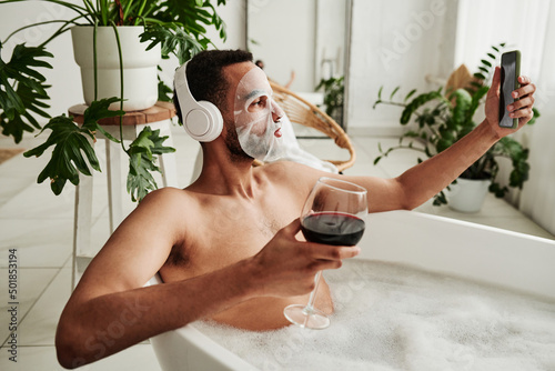 Foto Young man with facial mask drinking wine and making selfie portrait on mobile ph