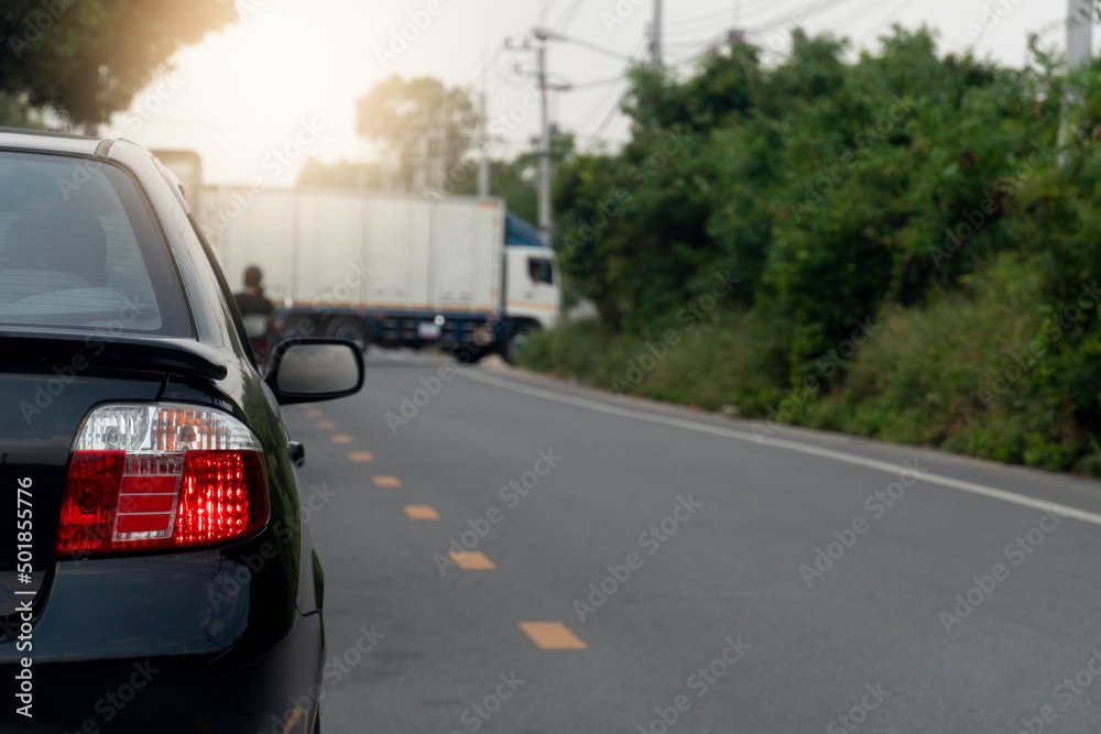 Rear of the black car turn on brake light for stop on asphalt road. Container trucks parked blocking traffic. Roads in provincial areas with trees on the side of the road.