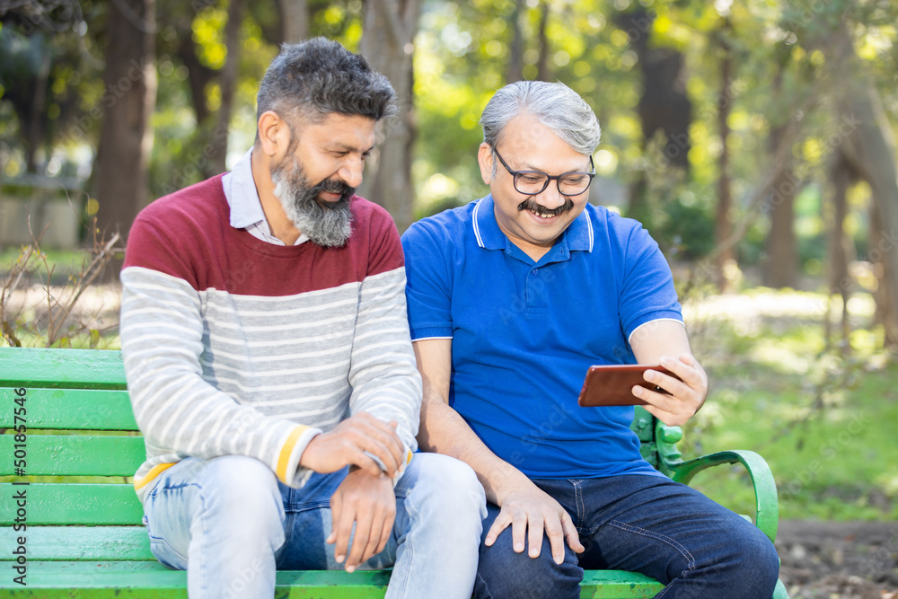 Two happy indian senior men watching video or social media on smart android phone while sitting at park outdoor, Mature old people using technology, Friends having fun. Two happy indian senior men watching video or social media on smart android phone while sitting at park outdoor, Mature old people using technology, Friends having fun.