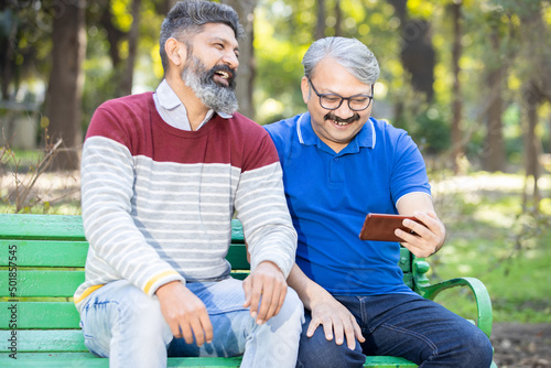 Two happy indian senior men watching video or social media on smart android phone while sitting at park outdoor, Mature old people using technology, Friends having fun. © gajendra