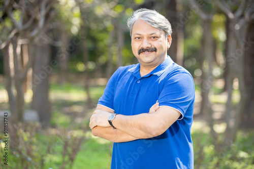 Fototapeta Naklejka Na Ścianę i Meble -  Happy senior indian asian man with arms crossed standing at park outdoor, Old mustache and grey haired male smiling wearing blue t-shirt. Copy space.