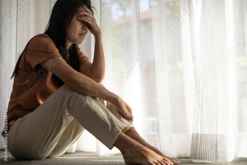 Unhappy woman sitting on the carpet and holding her forehead while having headache. Young asian woman with depression sitting alone in bed room. major depressive disorder failure sadness concepts