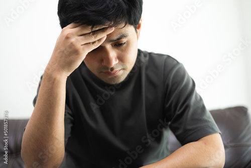 Unhappy handsome man sitting on the sofa and holding his forehead while having headache. Young asian man with depression sitting alone in bed room. major depressive disorder failure, sadness concepts photo