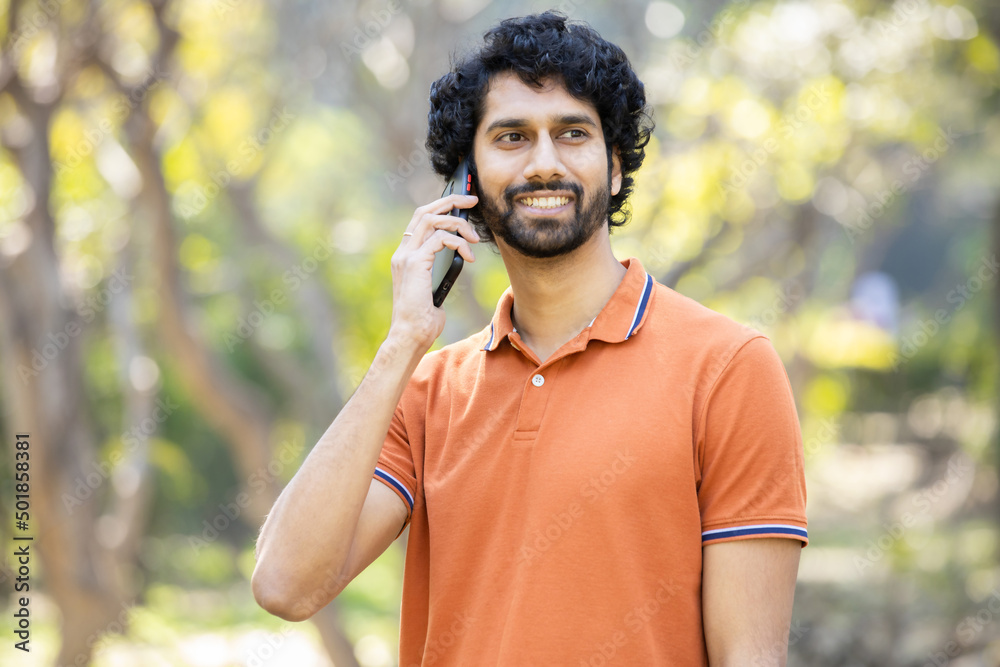 Obraz premium Young happy Indian man with beard and orange t-shirt talking on smart phone while standing in a park outdoor summer, Smiling male calling on mobile phone, technology and communication,Closeup.