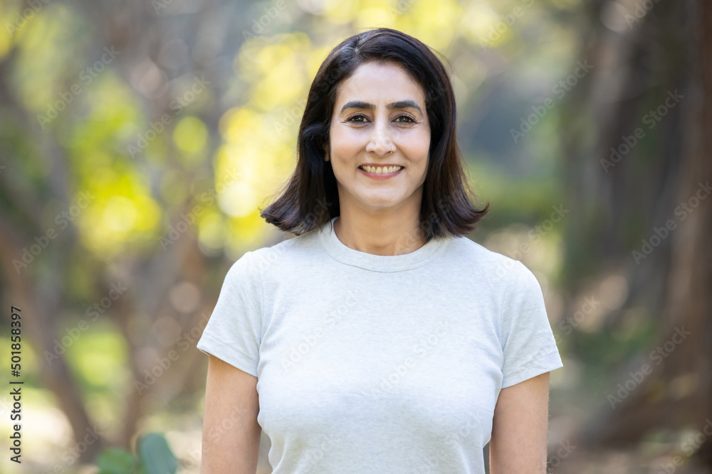 Smiling mature urban indian woman wearing white e t-shirt standing in the park outdoors, asian female with positive expression posing. closeup, copy space. Smiling mature urban indian woman wearing white e t-shirt standing in the park outdoors, asian female with positive expression posing. closeup, copy space.