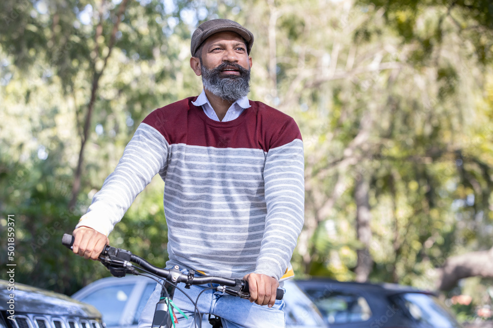 Happy Indian mature beard man wearing cap ride bicycle outdoor in the park outdoor.