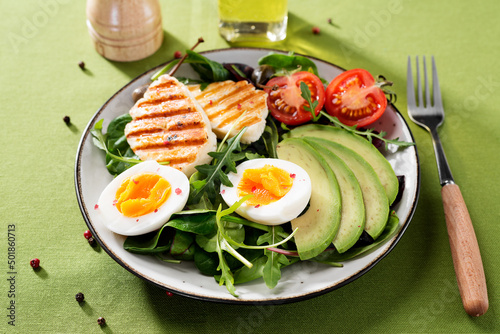 Healthy keto diet breakfast: boiled egg, avocado slices, grilled halloumi cheese, salad leaves. Green background.