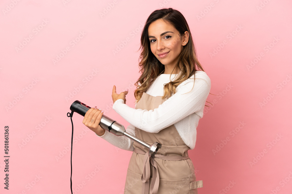 Young woman using hand blender over isolated pink background pointing back Young woman using hand blender over isolated pink background pointing back