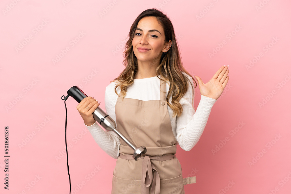 Young woman using hand blender over isolated pink background saluting with hand with happy expression Young woman using hand blender over isolated pink background saluting with hand with happy expression