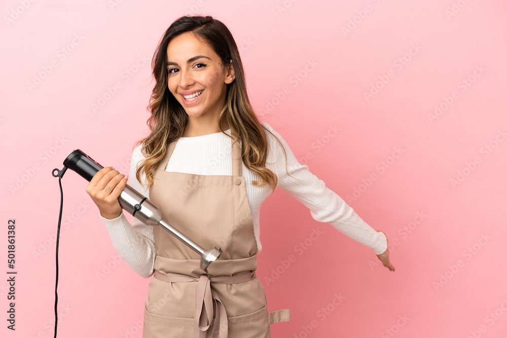 Young woman using hand blender over isolated pink background extending hands to the side for inviting to come Young woman using hand blender over isolated pink background extending hands to the side for inviting to come