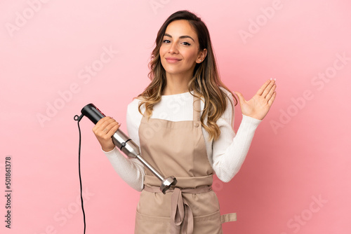 Young woman using hand blender over isolated pink background saluting with hand with happy expression
