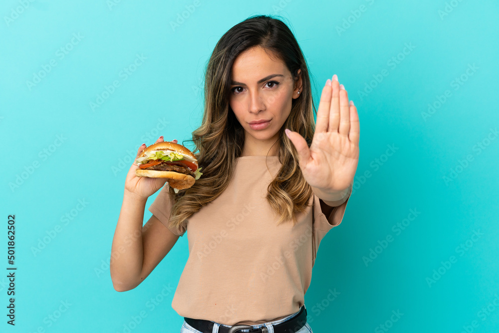Young woman holding a burger over isolated background making stop gesture Young woman holding a burger over isolated background making stop gesture