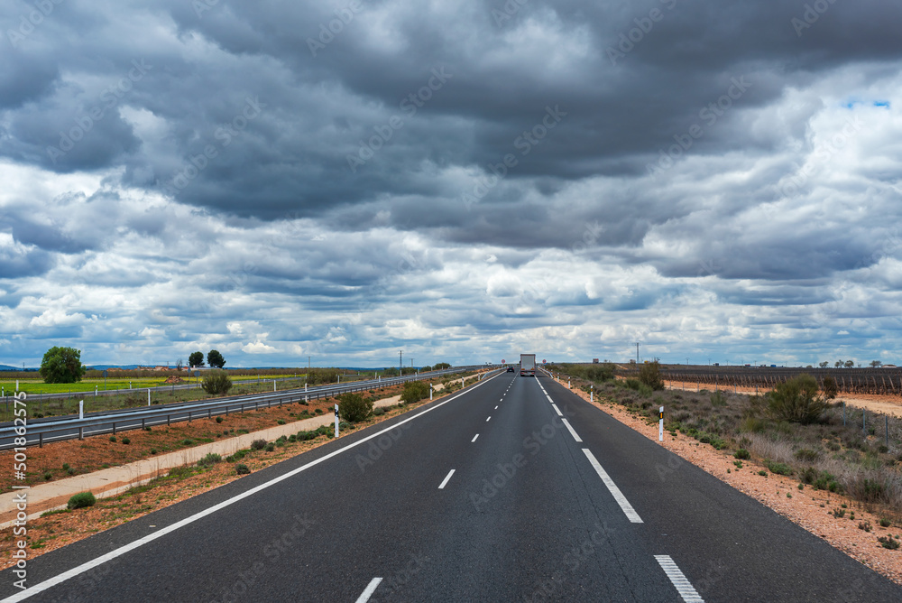 Fototapeta premium Truck driving along a straight highway with a flat horizon and a cloudy sky in the community of La Mancha, Spain.