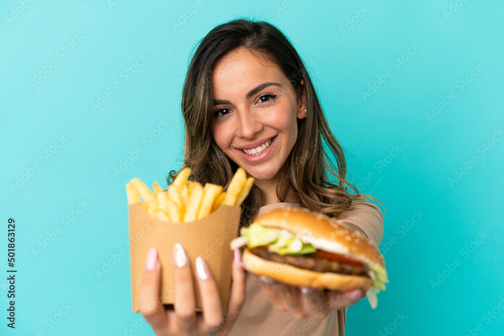 Young woman holding fried chips and burger over isolated background Young woman holding fried chips and burger over isolated background