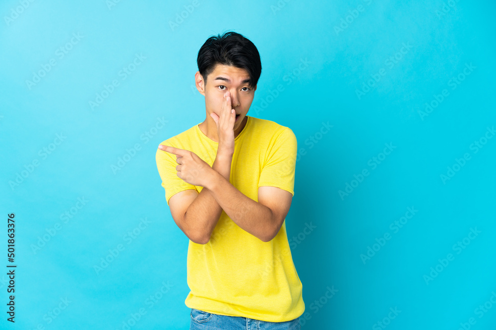 Young Chinese man isolated on blue background pointing to the side to present a product and whispering something Young Chinese man isolated on blue background pointing to the side to present a product and whispering something