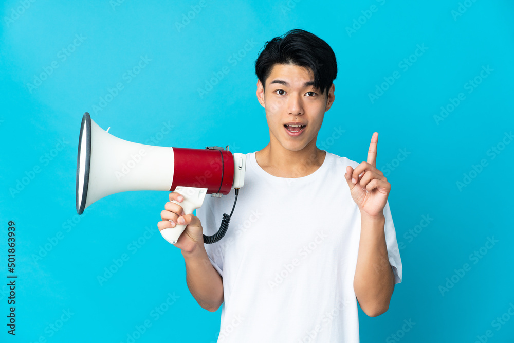 Young Chinese man isolated on blue background holding a megaphone and pointing up a great idea Young Chinese man isolated on blue background holding a megaphone and pointing up a great idea