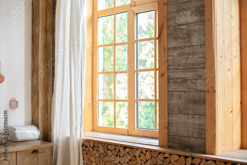 Big wooden window with frame and window sill and nature on background. Empty room, wooden window with with White linen curtain and logs decoration wall on a sunny day indoor shot. Scandinavian room