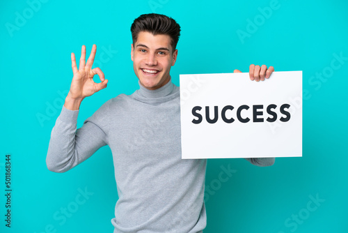 Young handsome caucasian man isolated on blue bakcground holding a placard with text SUCCESS and celebrating a victory © luismolinero