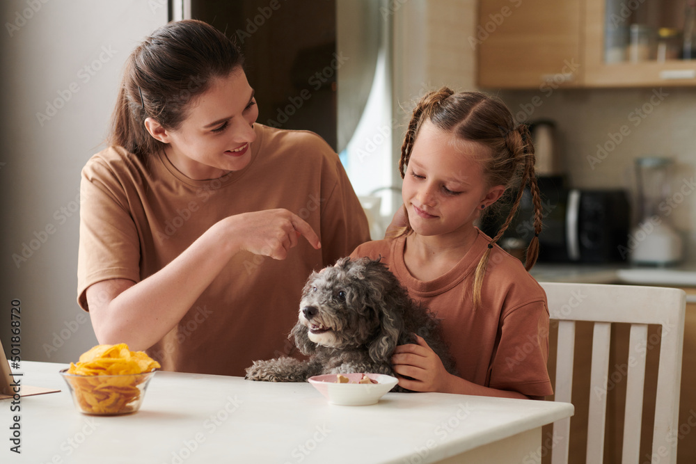 Smiling mother and daughter letting dog to eat from bowl on kitchen