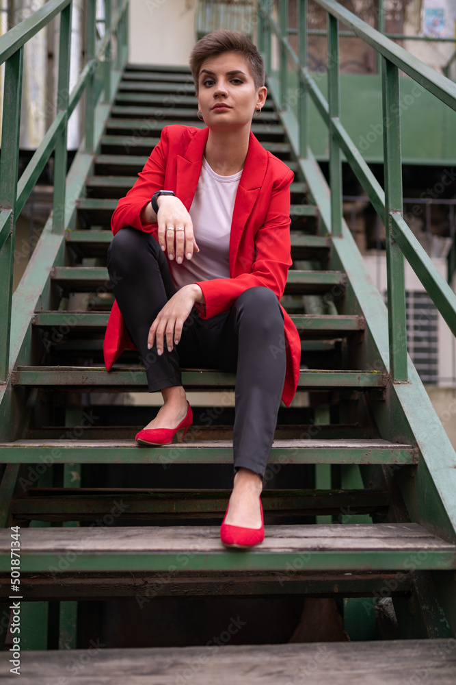 A stylish model girl with short brown hair in a red jacket and pumps, a white T-shirt, posing for a photographer sitting on a wooden staircase with wooden railings