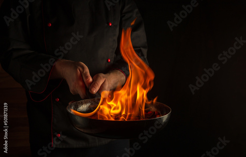 Professional chef cooking food in pan with fire flame on dark background. Restaurant and hotel service concept