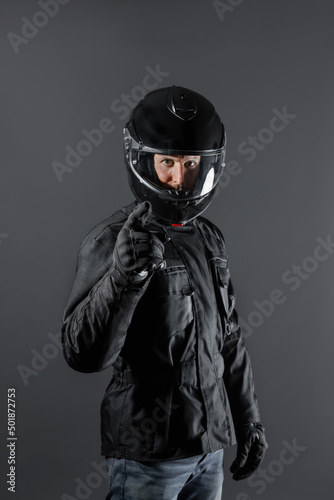 Studio shot of Motorcyclist biker in black equipment points and looks at camera.