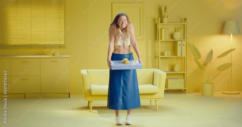 Full length of a happy blonde hair woman in sleeveless clothes smiling at the camera while holding a gift box. Carefree young female model getting a surprise present, standing on a yellow studio Full length of a happy blonde hair woman in sleeveless clothes smiling at the camera while holding a gift box. Carefree young female model getting a surprise present, standing on a yellow studio