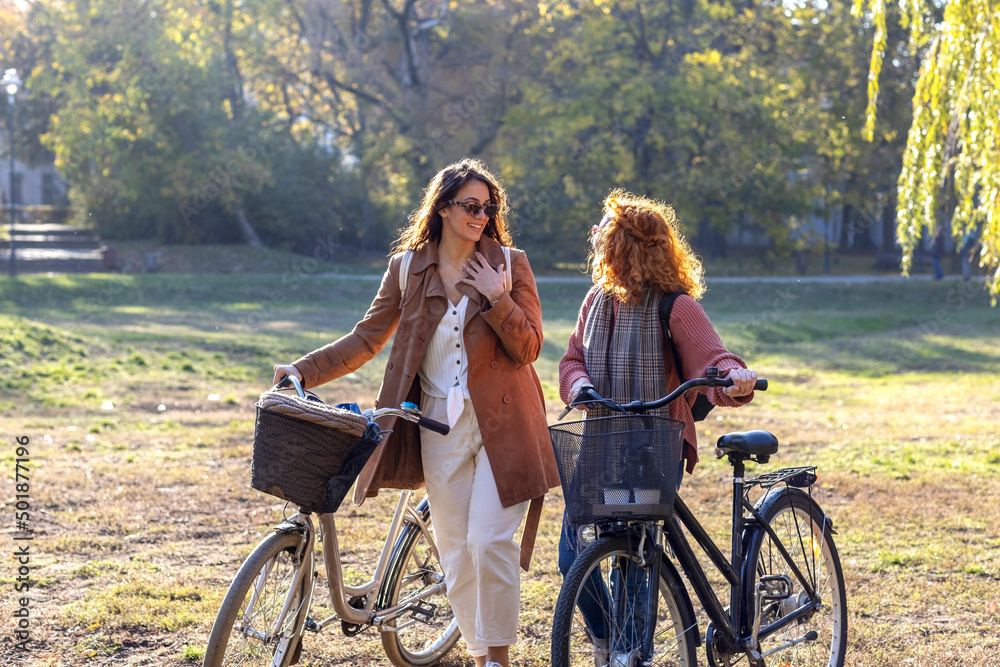 Fototapeta premium Two young woman with bikes walk and talk and smile in public park