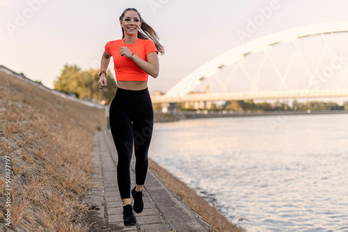 Young happy focused fitness girl in black yoga pants and orange short shirt jogs on riverbank during the day. Front view. photo
