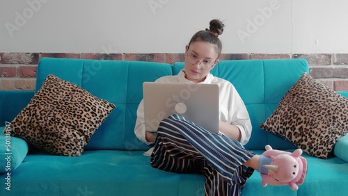 A young freelance woman in a shirt and slippers, working remotely in a home office, uses a laptop while sitting on the couch. A modern woman communicates in an online conference using a computer.