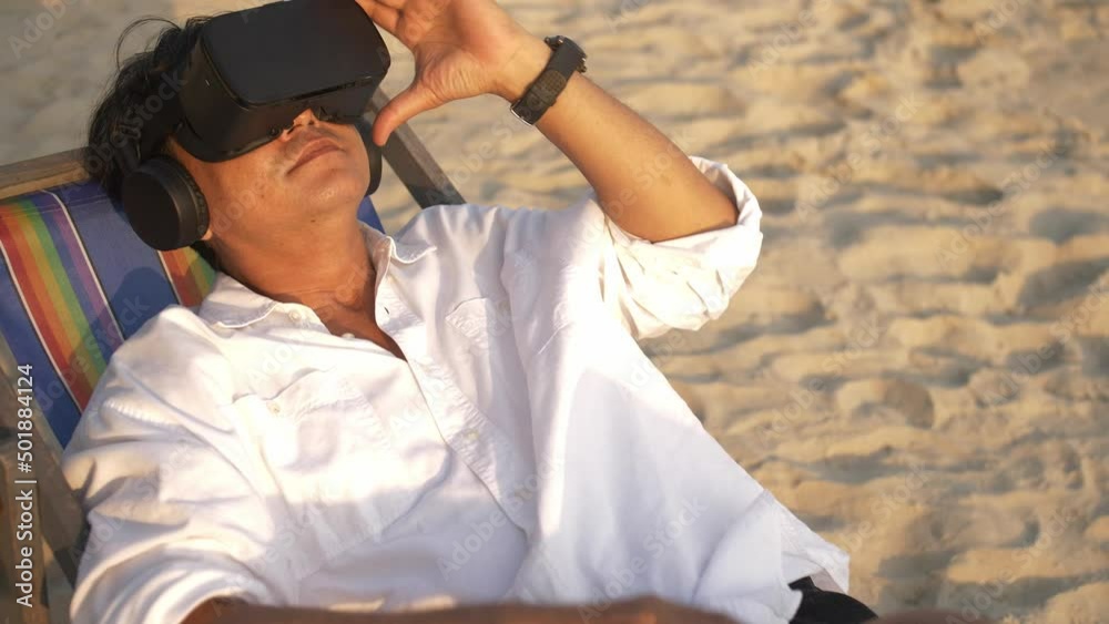 Asian man wearing virtual reality glasses while relaxing on the beach. Asian man wearing virtual reality glasses while relaxing on the beach.