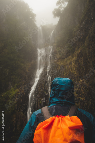 Rainy weather hiker in a waterproof jacket looks at the majestic Risco waterfall immersed in mist and rain on the island of Madeira, Portugal. Discovering magical places in Europe