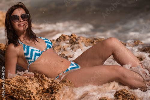 Young beautiful fit female model posing on the rocky stone in mini bikini while strong waves hitting the rocks on the background at Algarve beach Portugal 