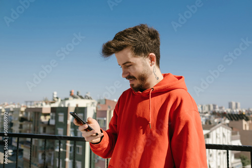 Handsome young man wearing red hoodie using phone. Man looking to smartphone screen with smiles. Messaging with friend or watching video or scrolling on social media.