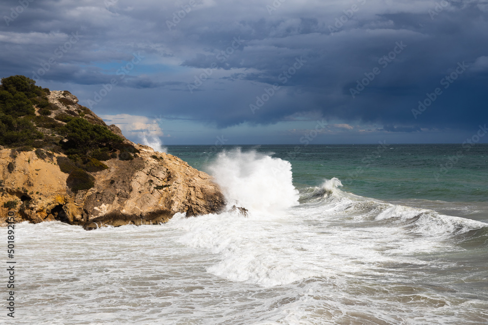 Stormy coastline with water splash and dark sky, sandy beach with turquoise sea, Sitges, Spain