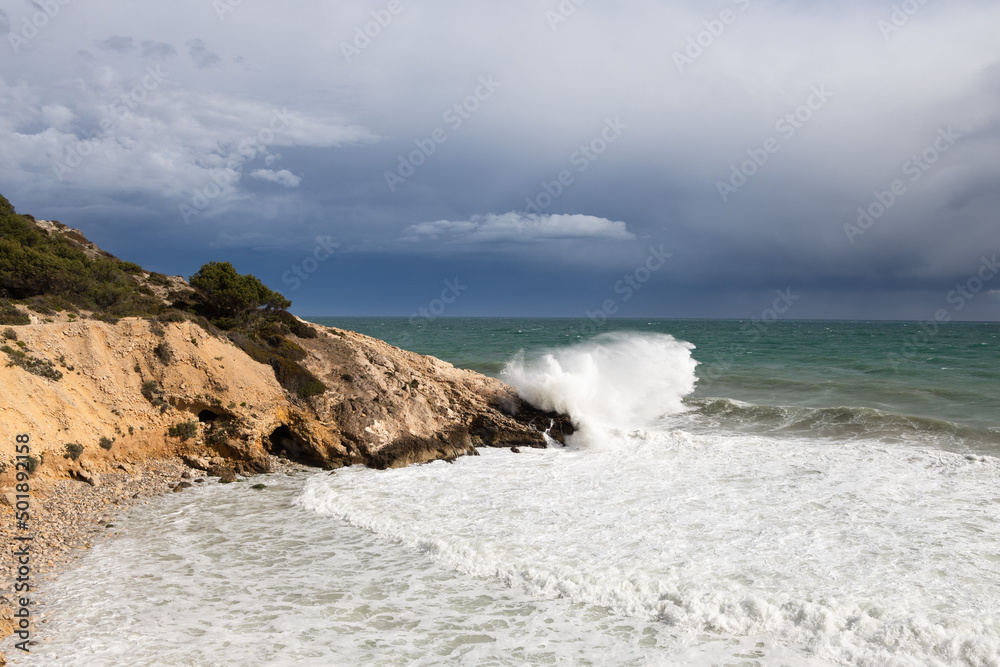 Stormy coastline with water splash and dark sky, sandy beach with turquoise sea, Sitges, Spain