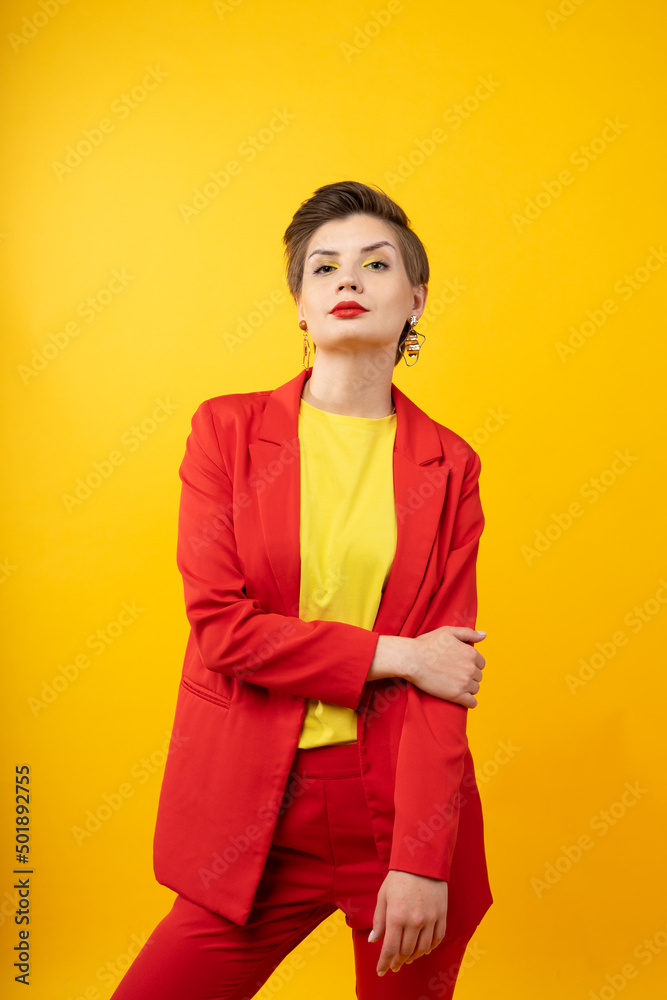 A woman in bright makeup, a red jacket, a yellow T-shirt and red trousers, with bright red lipstick and green shadows, stands on a yellow background with arm hold another elbow. Studio shooting A woman in bright makeup, a red jacket, a yellow T-shirt and red trousers, with bright red lipstick and green shadows, stands on a yellow background with arm hold another elbow. Studio shooting