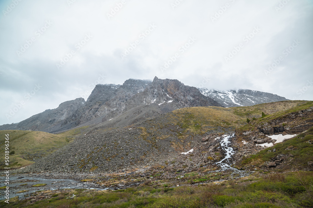 Atmospheric landscape with clear mountain stream from hills against snowy mountain range with peaked top in low clouds. Beautiful mountain creek and high sharp rocks under cloudy sky in gloomy weather
