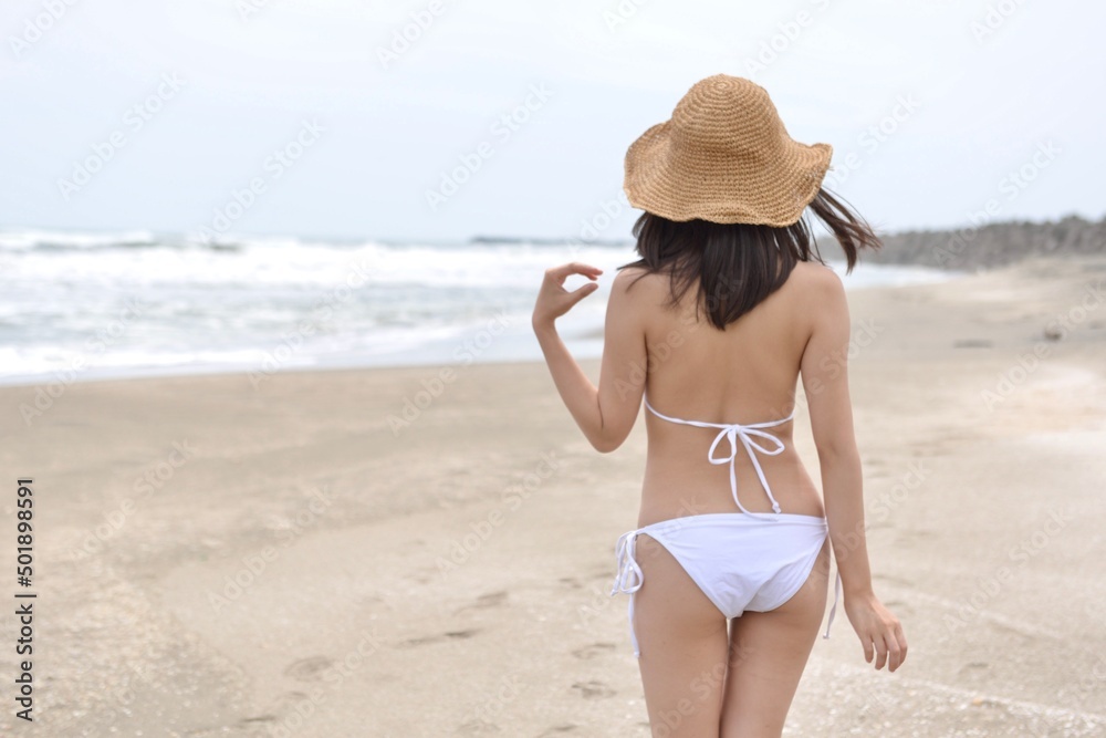 海岸でポーズをとる麦わら帽子を被った白いビキニの水着姿の若い女性の後ろ姿	