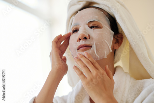 Young woman applying sheet mask on face to moisturize skin after shower