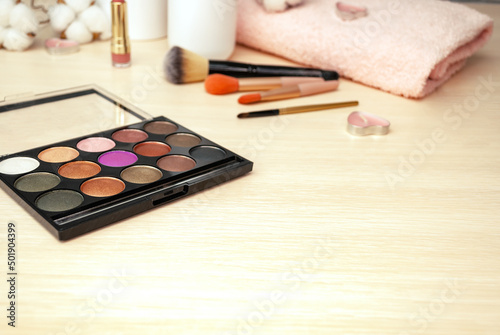 A palette with eye shadow, makeup brushes, towels, cotton flowers on a light wooden background. Morning face makeup. Selective focus. Copy space.