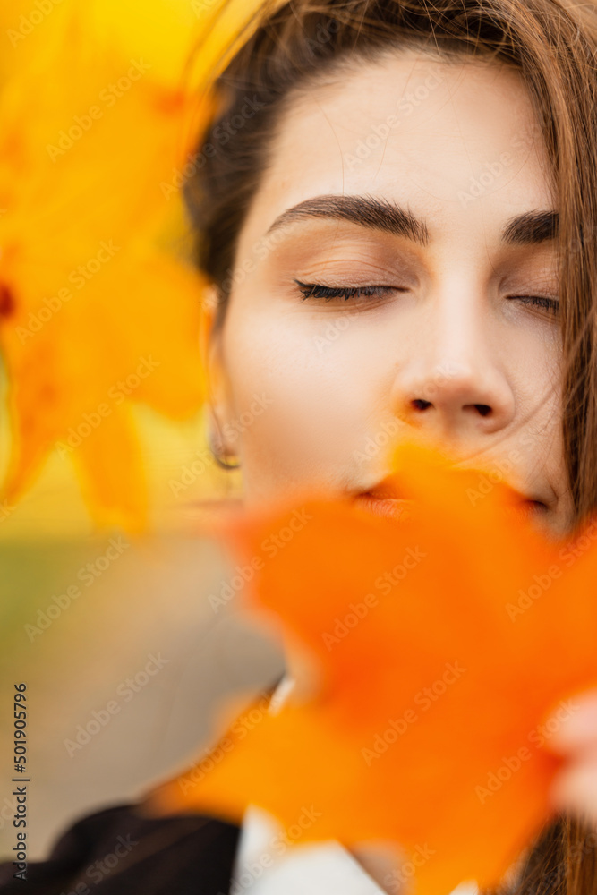 Fototapeta premium Autumn colorful portrait of a beautiful young gentle girl with a cute face and closed eyes in a golden fall park with bright yellow and orange foliage. Women's beauty and autumn leaves