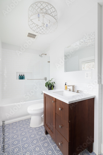 A renovated bathroom with a blue and white mosaic tile floor  wood vanity cabinet  white chandelier  and a subway tile shower.