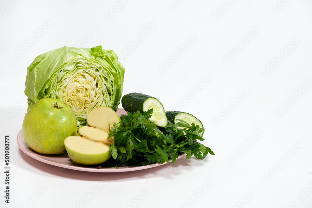A plate with green vegetables and fruits - cabbage, apples, cucumbers and parsley on a white background with a place for text A plate with green vegetables and fruits - cabbage, apples, cucumbers and parsley on a white background with a place for text
