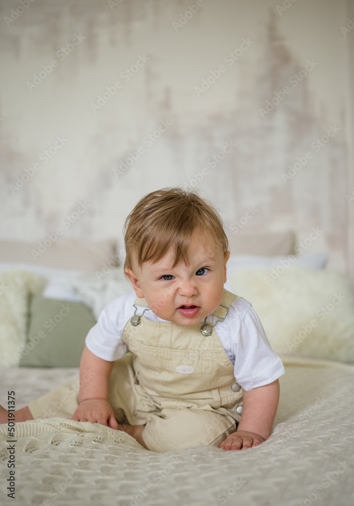 portrait of a funny little boy in a beige jumpsuit sitting on a bed in a room and making grimaces portrait of a funny little boy in a beige jumpsuit sitting on a bed in a room and making grimaces