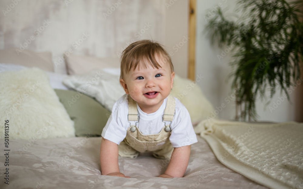 a cheerful little boy in a beige jumpsuit crawls on the bed in the room. Learn to crawl